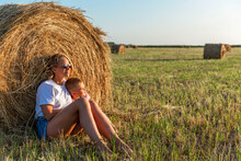 Mom And Her Little Son Are Sitting In A Field With Hay Rolls In The Setting Sun. A Woman In Denim Shorts And A White T-shirt. Farming And Nature.