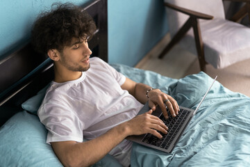 Job in bedroom, social networks, surfing, browsing on internet. Serious young man woke up, sits on bed, works on laptop in interior.