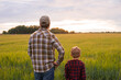 © Acronym - Farmer and his son in front of a sunset agricultural landscape. Man and a boy in a countryside field. Fatherhood, country life, farming and country lifestyle concept.