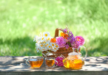 Wild Flowers In Basket, Glass Teapot And Cups With Herbal Tea On Table In Garden, Natural Background. Summer Season. Beautiful Rustic Composition. Relax Time. Useful Calming Tea. Tea Party Concept