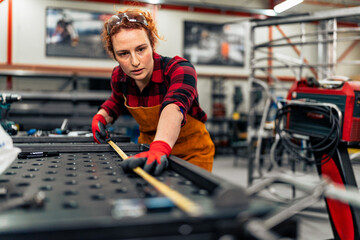 A young engineer measures and marks the measurements on a metal bar that he will test for durability, the workshop in the background and she is in protective equipment