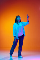 Young woman in casual clothes with raising hand and finger leading presentation against gradient orange studio background in neon light