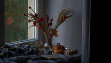 Beautiful Atmospheric Photograph Of Autumn Mood.Vase With Branches Of Orange Physalis, Pumpkins,light Garland,blanket And Book On Windowsill Near Window Wet From Rain.Autumn, Fall, Hygge Home Decor.