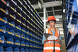 Young Asian female employee worker in safety vest and helmet using tablet checking products spare parts on shelf in industry factory warehouse