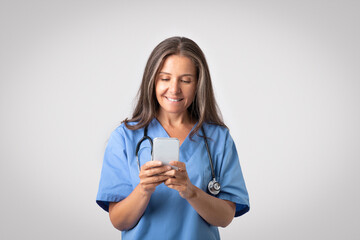 Professional senior female physician using smartphone with medical app, standing over light grey background, free space