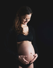 Wall Mural - Atmospheric shot of pregnant woman in underwear gently holding her naked round belly. Last month of pregnancy - 36th week. Side angle view. Black background.