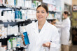 Young female pharmacist in medical uniform posing with cosmetic product in her hands in pharmacy