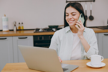 Young housewife latin IT woman wear casual clothes striped shirt hold use work on laptop pc computer talk mobile cell phone sit at table in light kitchen at home alone. Lifestyle cooking food concept.