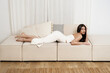 Beautiful young woman lying in a trendy white dress on sofa, indoors.