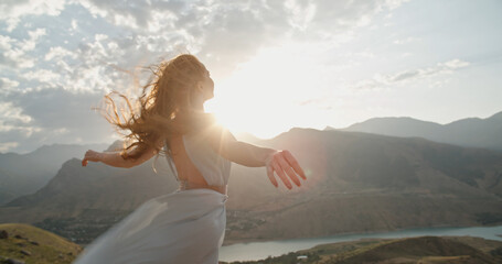 Woman in white dress standing on top of a mountain at sunset with raised hands while wind is blowing her dress and red hair - freedom, nature concept. Copy space
