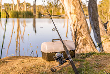A Fishing Road And Tackle Box Sitting At The Base Of A Gum Tree On A River Bank