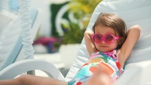 Funny cute baby girl on summer vacation. Child having fun in swimming pool. Sweet toddler girl in colorful swimsuit and sunglasses relaxing on sunbed.
