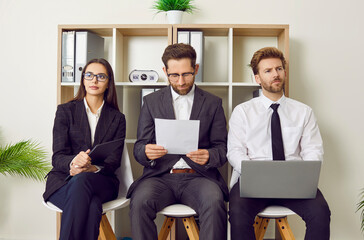 Canvas Print - Young business people sitting on the chairs in a row with resumes and laptops in hands. Group of a staff and company employees at work. Job candidates seekers waiting for interview invitation turn.