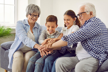 Wall Mural - Happy grandparents and little children having fun while spending time together. Cheerful, joyful brother and sister together with grandmother and grandfather sitting on sofa at home and stacking hands