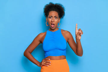 Young black woman in blue top and orange skirt posing against blue wall, holding one hand on waist, index finger of another hand is up, business time concept, copy space