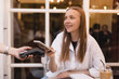© zvkate - Blonde attractive woman paying with cellphone resting in cafe while waitress holding payment terminal. Girl relaxing in restaurant. Focus on part payment terminal and mobile phone.