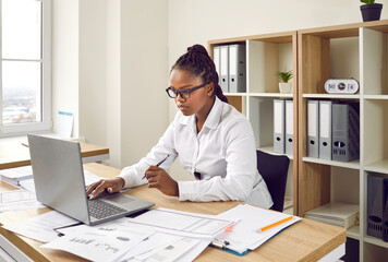 Wall Mural - Businesswoman working in her office. Young African American woman in a white shirt and glasses sitting at her desk, using a modern laptop computer, doing paperwork, working with business documents