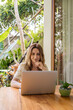 © Julija - Young beautiful woman sitting in a garden with laptop in a tropical resort. Girl with laptop computer connecting to 4G wireless for browsing social media working remotely at terrace