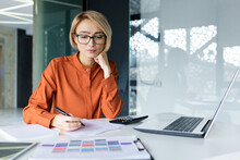 Serious And Pensive Business Woman Behind Paper Work Inside Office, Female Financier Worker Thinks About Contracts And Reports With Charts And Graphs, Blonde Successful Woman Uses Laptop At Work.