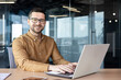 © Tetiana - Portrait of a young male student confidently sitting at a desk in the office and smiling at the camera, learning remotely from a laptop