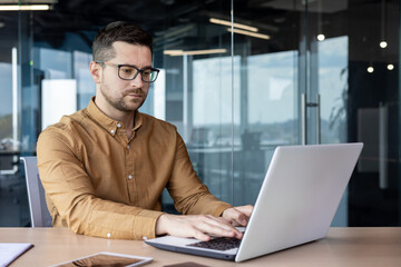 Portrait of a serious young male programmer, specialist developer who is concentrating on working on a laptop while sitting at a desk in an office center