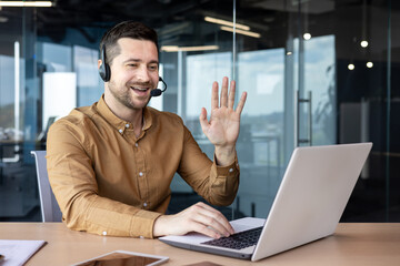 A young businessman in a headset smilingly talks on a video call on a laptop. Sitting in the office at the table and holding a business meeting, training, greeting and waving at the camera