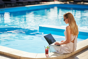 Self employed woman doing remote work with laptop sitting near swimming pool at tropical spa with cocktail drink. Freelancer businesswoman telecommuting with team on project from luxury resort.