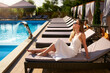 © artiemedvedev - Pretty woman sunbathing and chilling near swimming pool on beach bed at tropical resort. Attractive girl in white dress and sunglasses lies in the sun on deck chair in spa, relaxing on hot summer day.