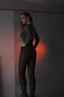 © NASTYA PALEHINA - young teen girl in a shiny silver bodysuit is standing fashion from the back with hands near waist and looking away on gray wall background in loft interior with red light. fashion concept, free space