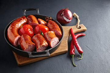 Wall Mural - sliced fried pork sausage in a cast iron pan with tomatoes on a wooden cutting board on a black stone table