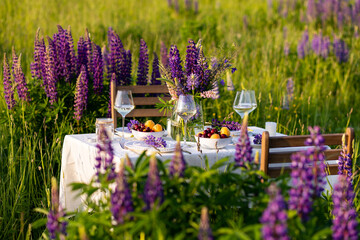 Beautiful countryside wedding of romantic dinner decor on a blooming meadow with purple violet lupins flowers. Vase, wineglasses, cherry and apricot. Wooden table, basket. Sunset, golden hour, summer