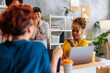 © bernardbodo - African-American business woman talking with colleague in front of two other coworkers.