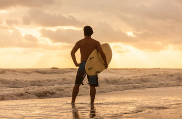 Rear view of young man male surfer with white surfboard looking at surf on a beach at sunset or sunrise