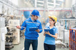 © Nutchapong Wuttisak - An American male engineer manager and female employee discuss business systems and projects in a large plastic and steel factory. Helmets, uniforms and listnotes, walkie-talkies, machinery working.