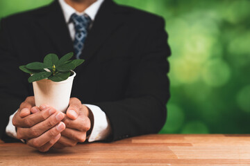 Businessman holding plant pot with his hand promoting forest regeneration and natural awareness. Ethical green business with eco-friendly policy utilizing renewable energy to preserve ecology. Alter