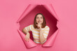 © sementsova321 - Beautiful brunette woman wearing striped top looking through ripped pink paper hole keeps finger near lips keeps secret showing shh gesture to camera.
