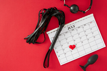 Wall Mural - Theme of BDSM Day for intimate exploration. Top view arrangement of calendar marking special date, anal plug, gag, leather whip, heart on red background with blank space for text or promotion