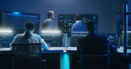 Multiethnic team of IT technical specialists and software programmers work with data server, analysis charts and blockchain network on computers and big digital screens in monitoring office at night.