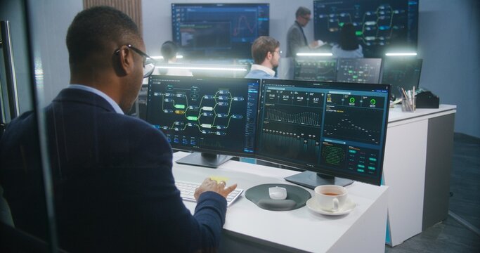 African American big data scientist works at computer in monitoring room. Multiracial colleagues analyze real-time charts at background. Multiple big screens on the wall with displayed server data.