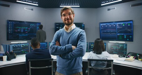 Adult IT scientist smiles, stands in monitoring room and looks at camera. Big data engineers work at computers at background. Multiple big screens with displayed real-time analysis charts. Dolly shot.