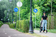 Young blonde hair girl with stroller walking near the bike road with blue road sign or signal of bicycle lane among green trees and spring or summer nature and street lamps background