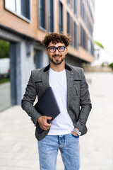 Wall Mural - Handsome young businessman holding laptop while using his phone and standing on the street