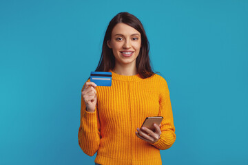 Portrait of a smiling casual girl in orange sweater holding mobile phone and plastic credit card isolated over blue background