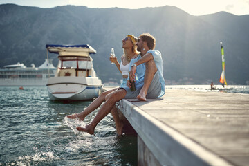 Wall Mural - Couple having a drink and laughing by seaside sitting on wooden jetty by water. Couple in love. Holiday, lifestyle, togetherness concept.