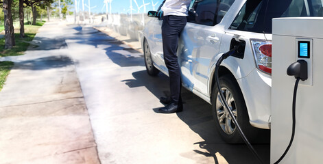 charging electric vinhicle on ev station ner public park and  wind turbine and man near car breesing clear air