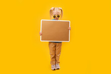 Child Holds Blank Cork Board. Blonde Girl In Sunglasses Holds Board With Space For Your Text. Portrait Of Small Girl On Yellow Background.