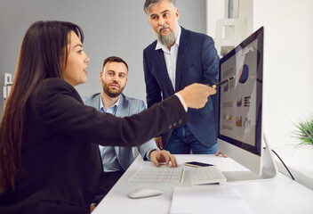 Young attractive business woman showing her colleagues stats financial data on pc computer screen with diagram, charts sitting at the desk in office analyzing company growth on meeting.