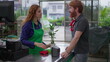 © Marco - Male customer buying plant at horticulture cashier. Female employee wearing green apron interacting at checkout with client at local business store