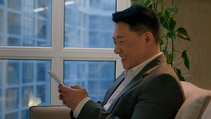 Middle-aged Asian businessman happy smiling multiracial chinese man browsing internet social media on mobile phone in office joyful male entrepreneur use cellphone gadget business district window view