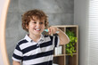 Cute little boy brushing his teeth with electric toothbrush near mirror in bathroom, space for text